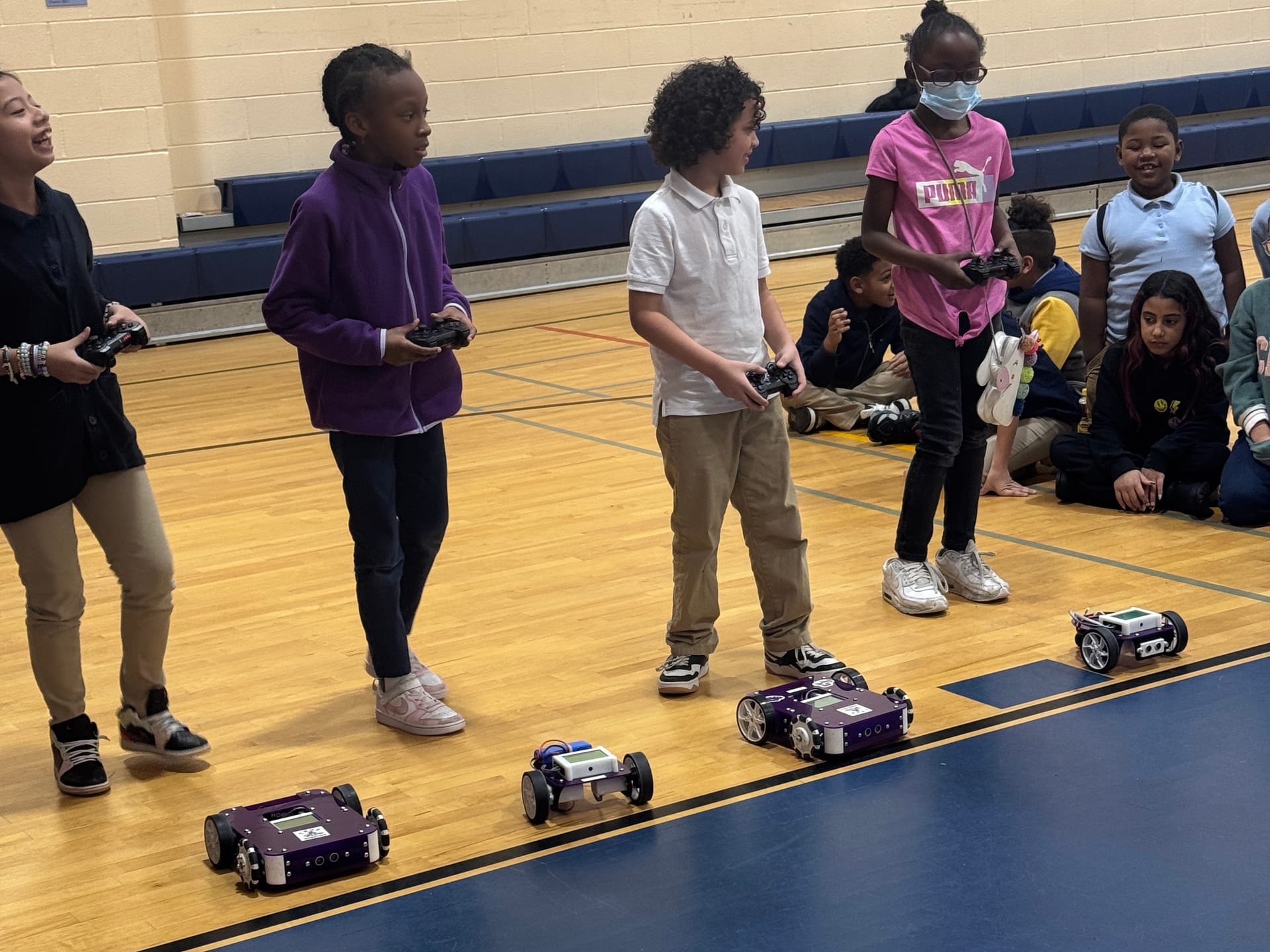 Four elementary students line up with game controllers to drive Axle and Pilot robots across a gym floor while classmates watch