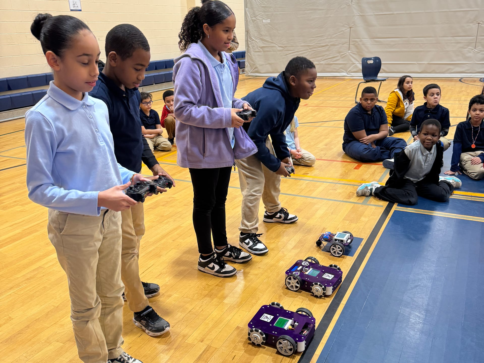 Four students stand in a row with game controllers while their purple robot kits drive down the gym floor in a line