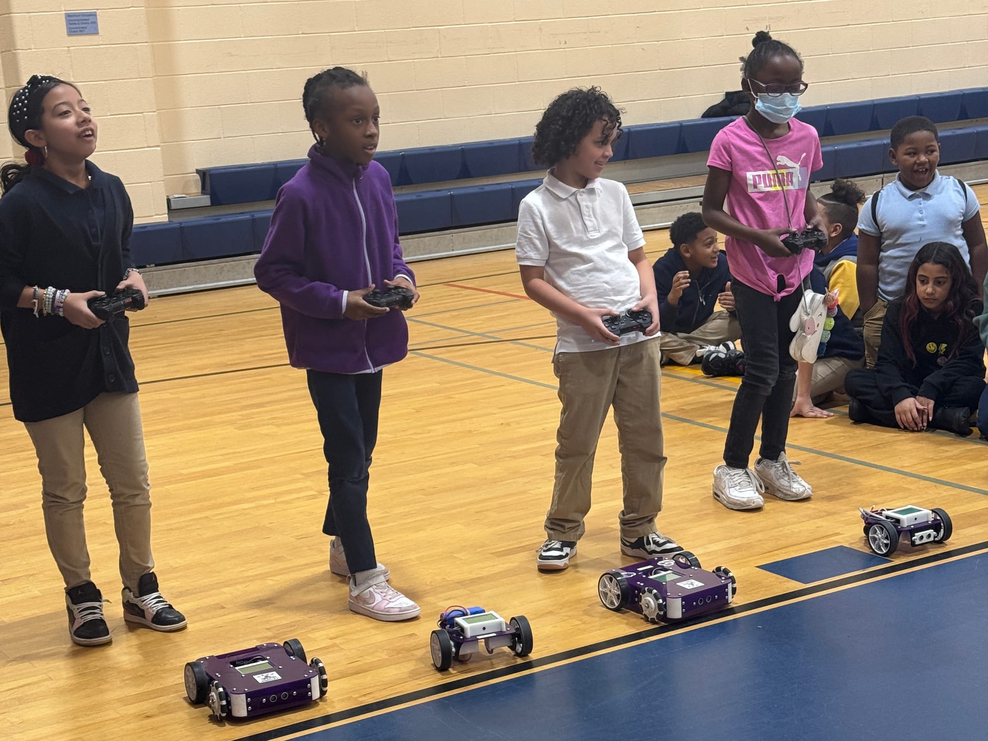 Four students hold game controllers in a school gym while their robot kits line up on the floor in front of them