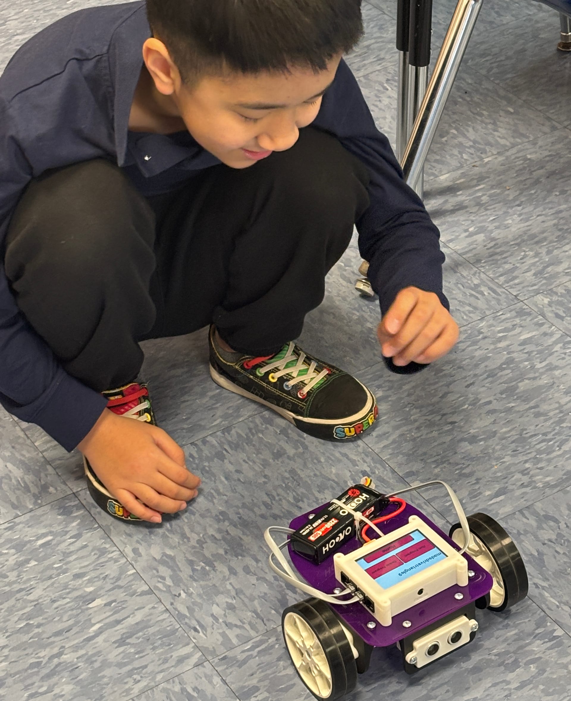An elementary student crouches on the classroom floor to watch a freshly built Pilot robot drive