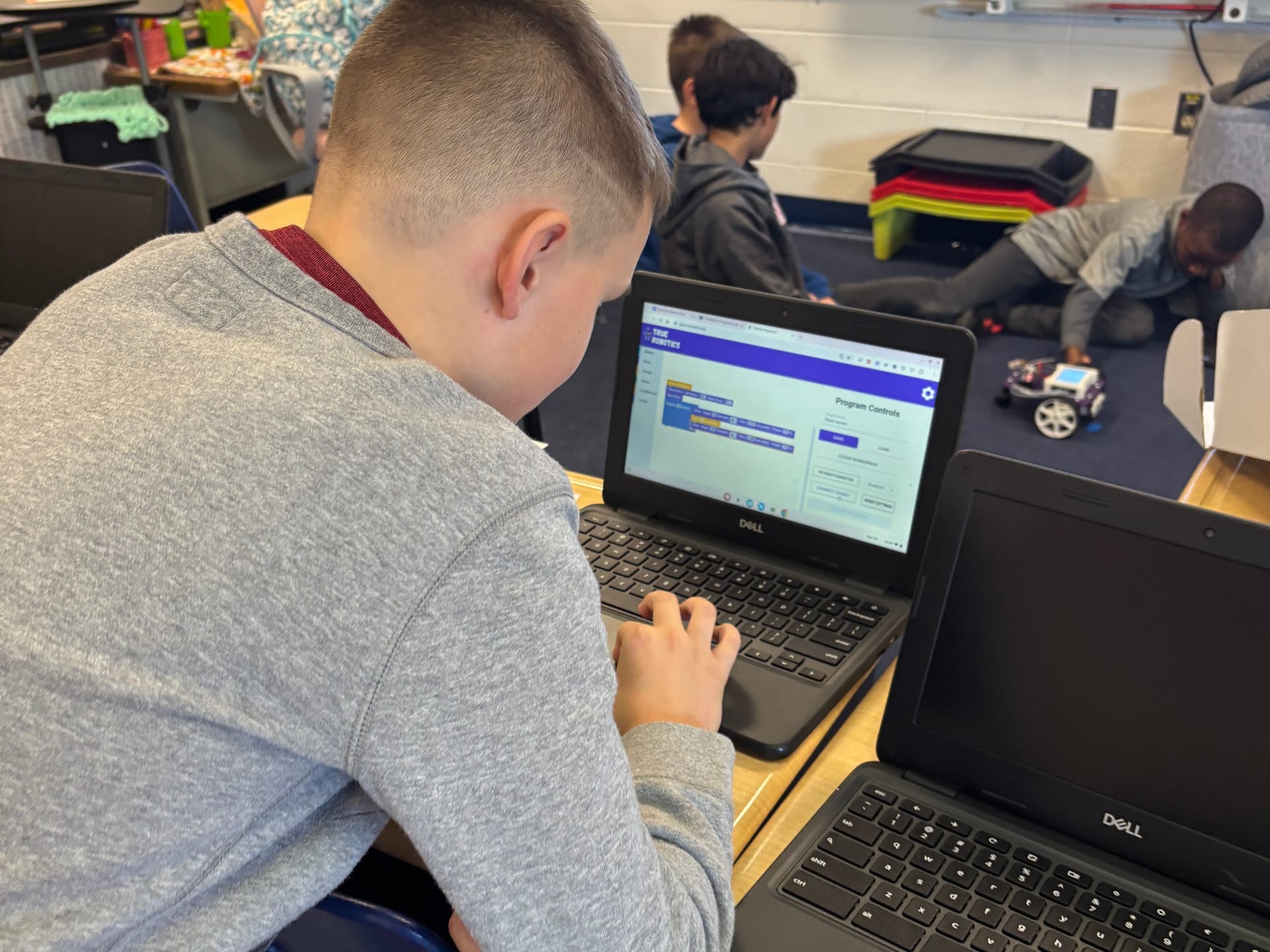 An elementary student programs a Pilot robot in True Robotics Playground on a Chromebook while classmates test theirs on the floor behind