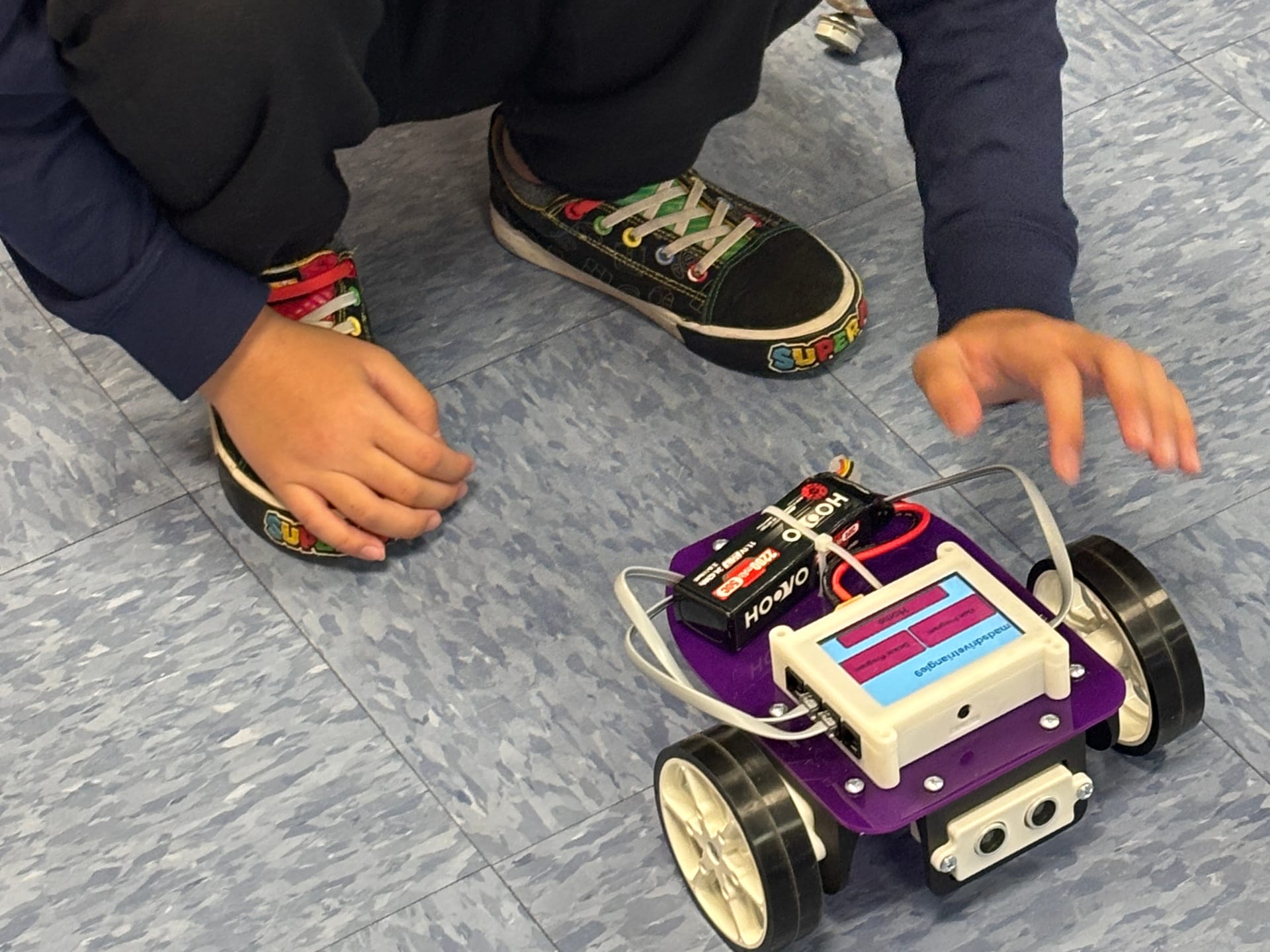 A student crouches on the classroom floor with both hands out to guide a purple Pilot robot across the tile