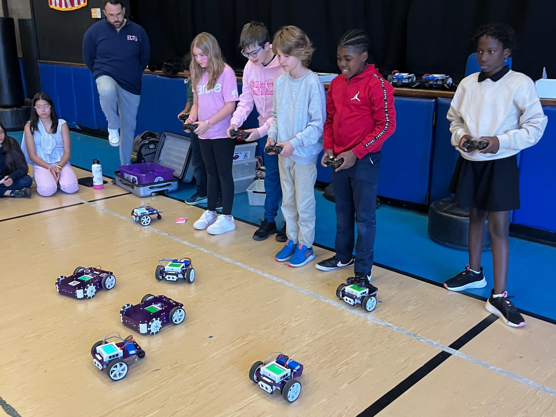 A row of students in a gym drive Axle robots with game controllers while a teacher looks on