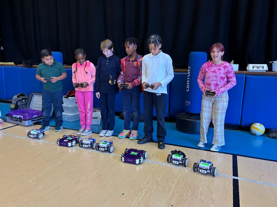 Six students stand in a row with game controllers driving a line of purple robot kits across a school stage