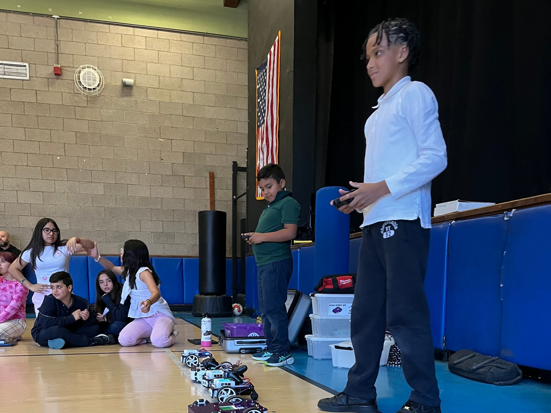 Two students hold game controllers beside a line of robot kits on a gym floor while classmates sit and watch