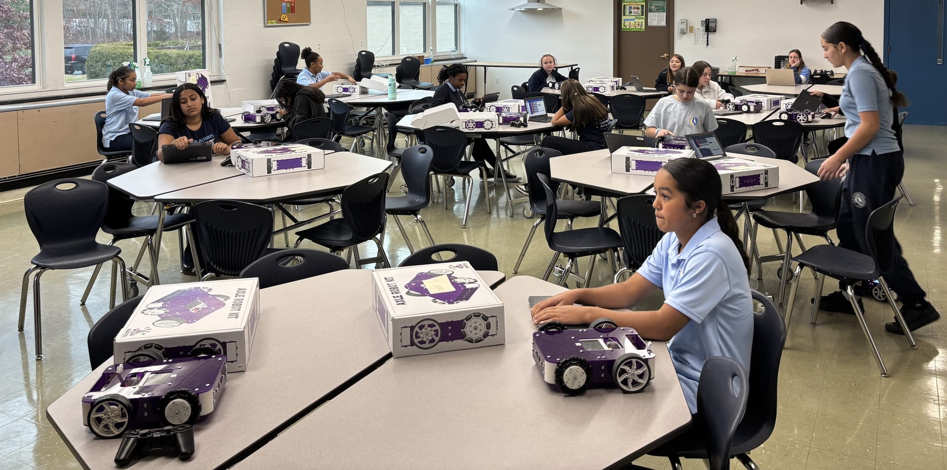 Middle school students at classroom tables open Axle Robot Kit boxes with a built robot in the foreground