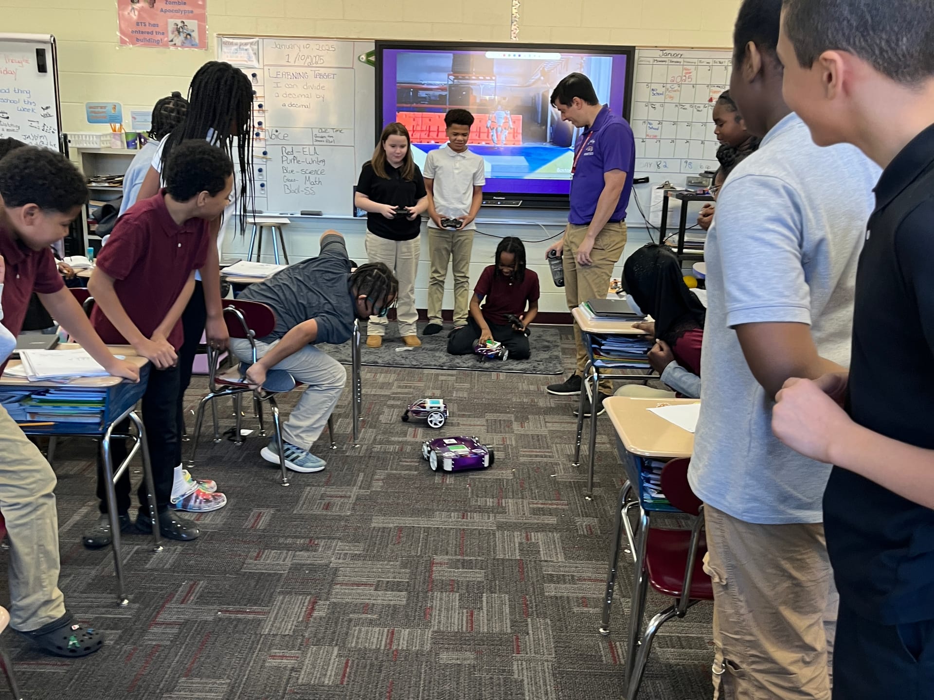 Middle school students gather along a classroom aisle to watch Axle and Pilot robots drive during a True Robotics demo