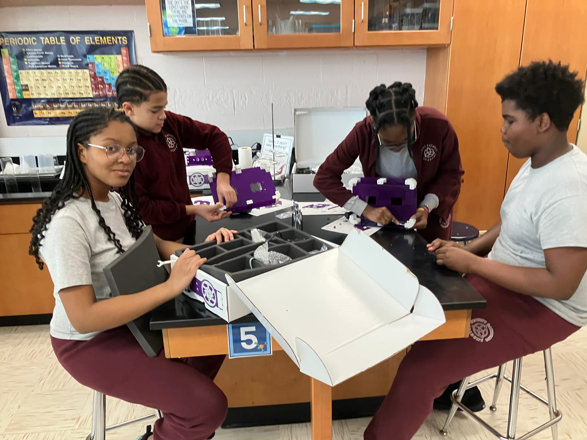 Three middle school students unpack a robot kit box together at a science lab table