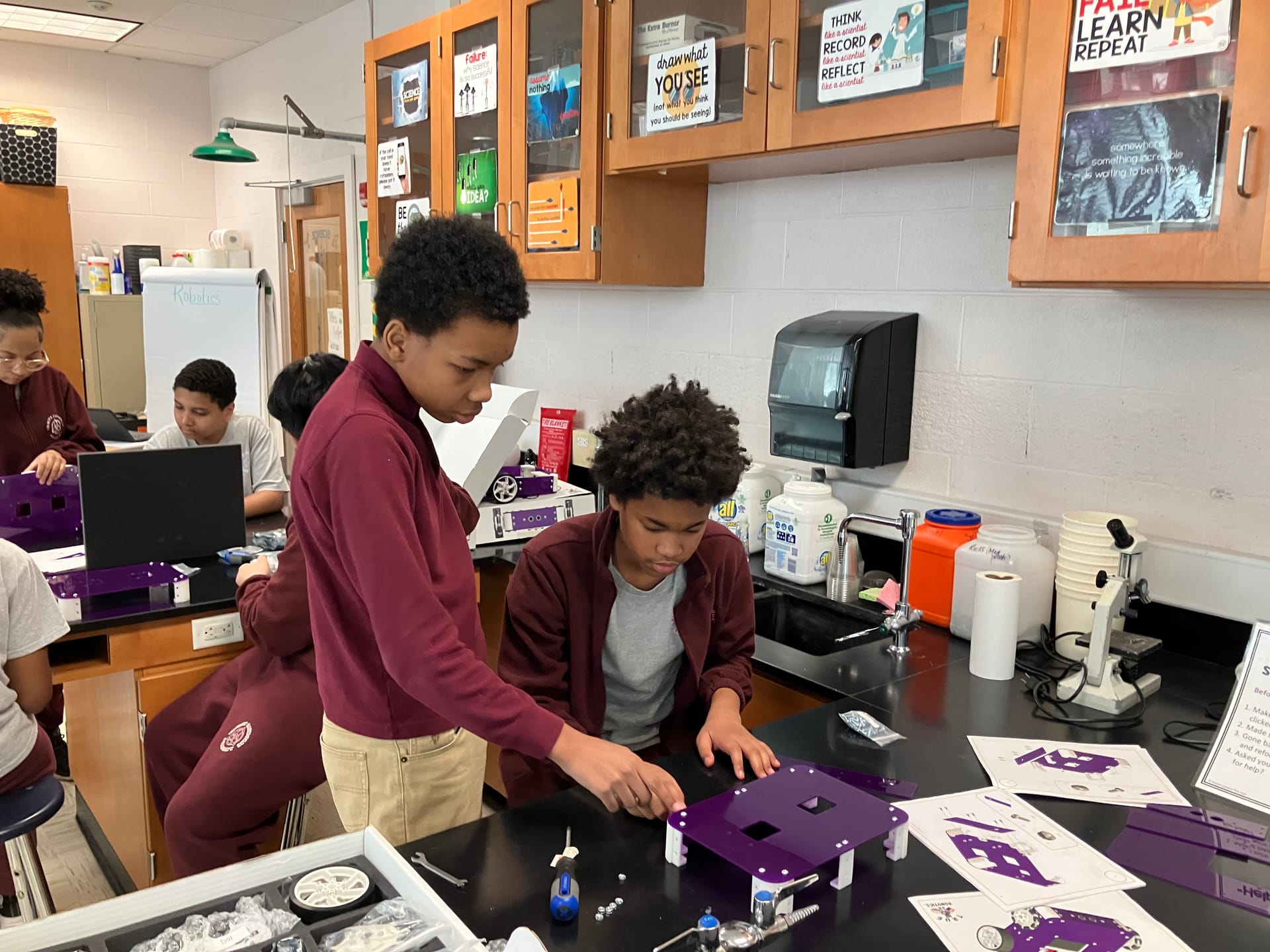 Two middle school students assemble a purple robot chassis at a lab bench with the build guide open beside it