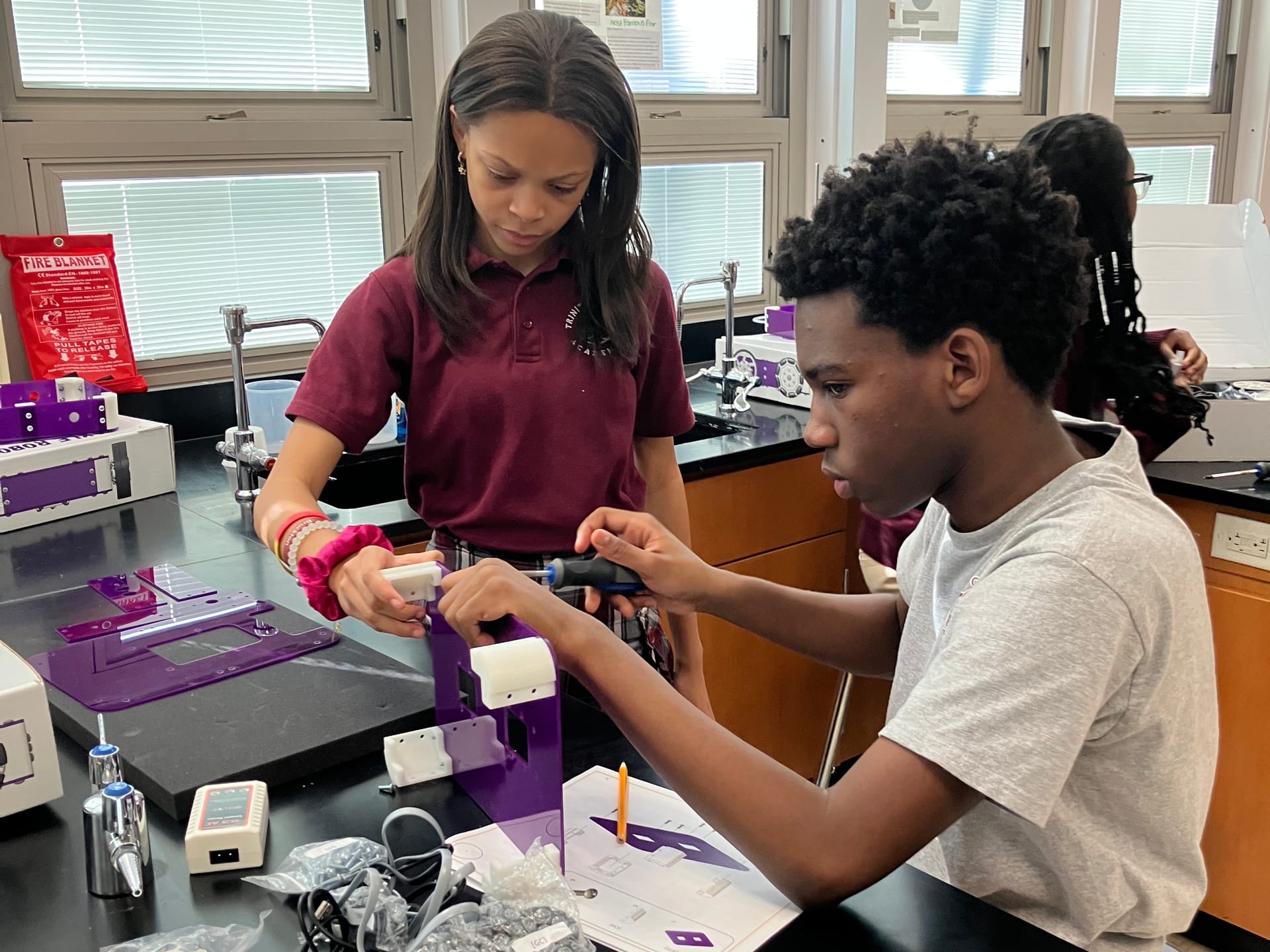 Two students at a science lab bench build a purple robot kit together, one turning a screwdriver while the other holds a part beside the printed guide