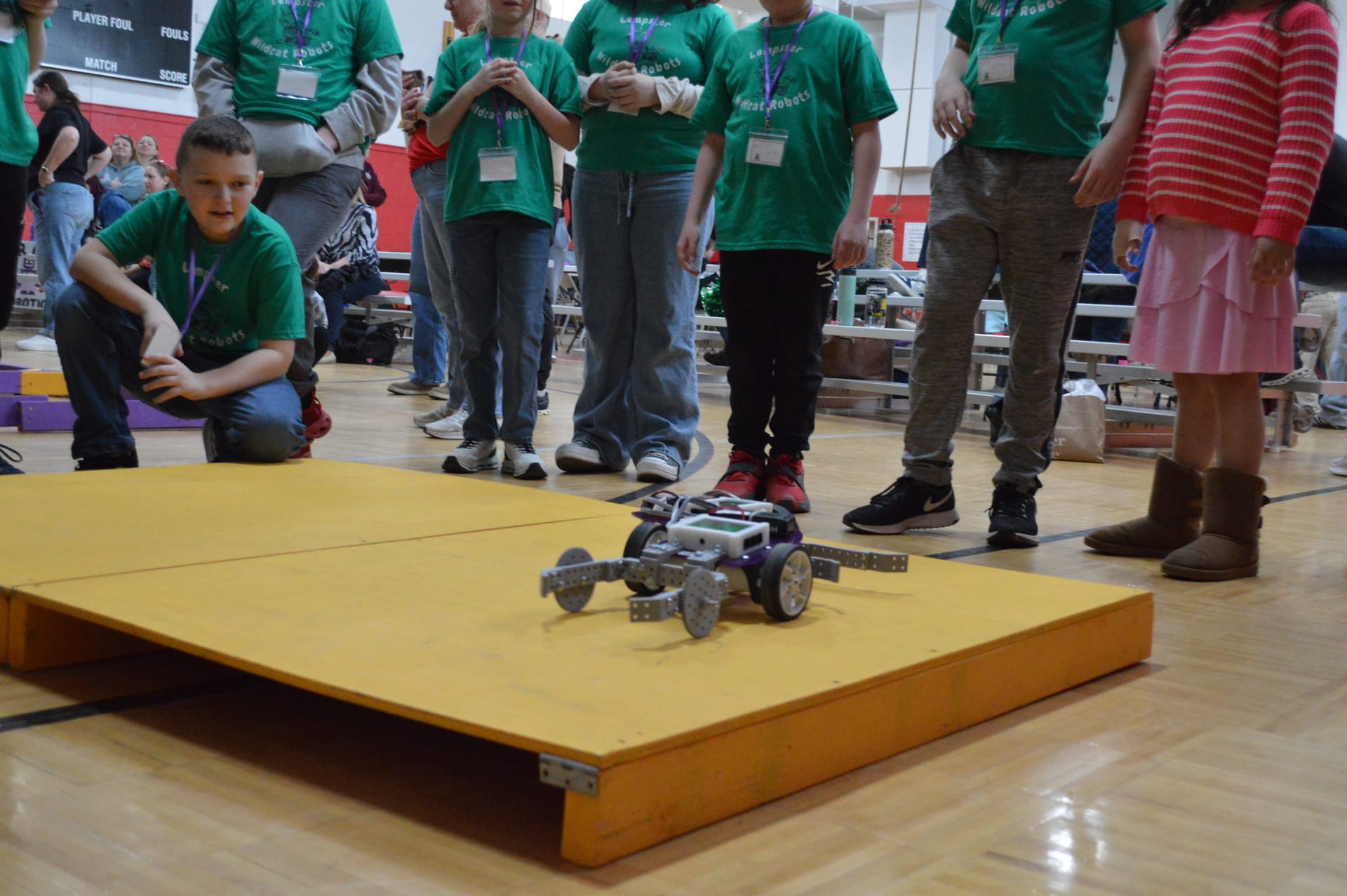 A Pilot robot with FlexForge arms climbs the yellow ramp while a team in green shirts watches from the sideline