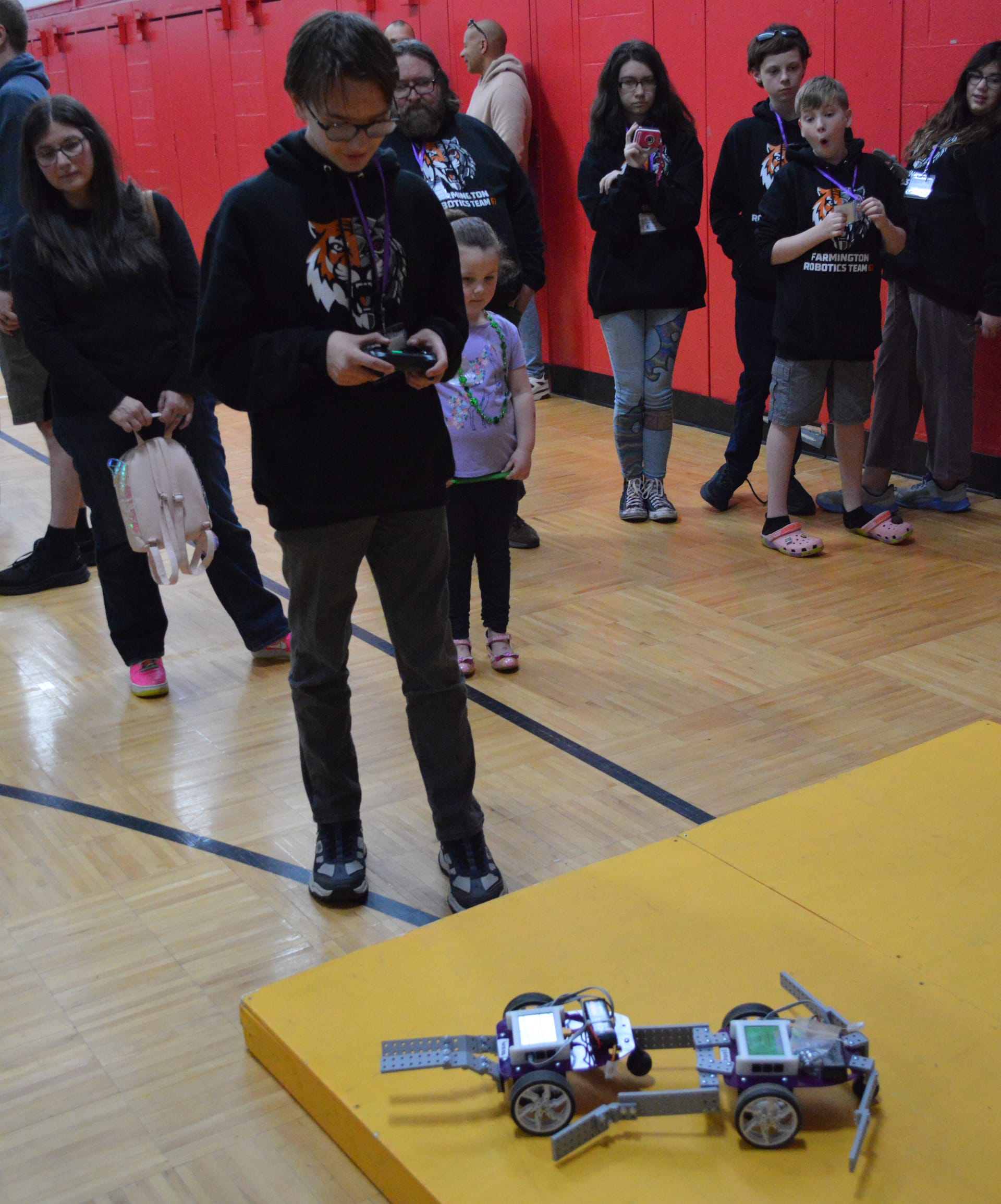 A student drives a Pilot robot with FlexForge attachments up the yellow ramp with a game controller while families watch