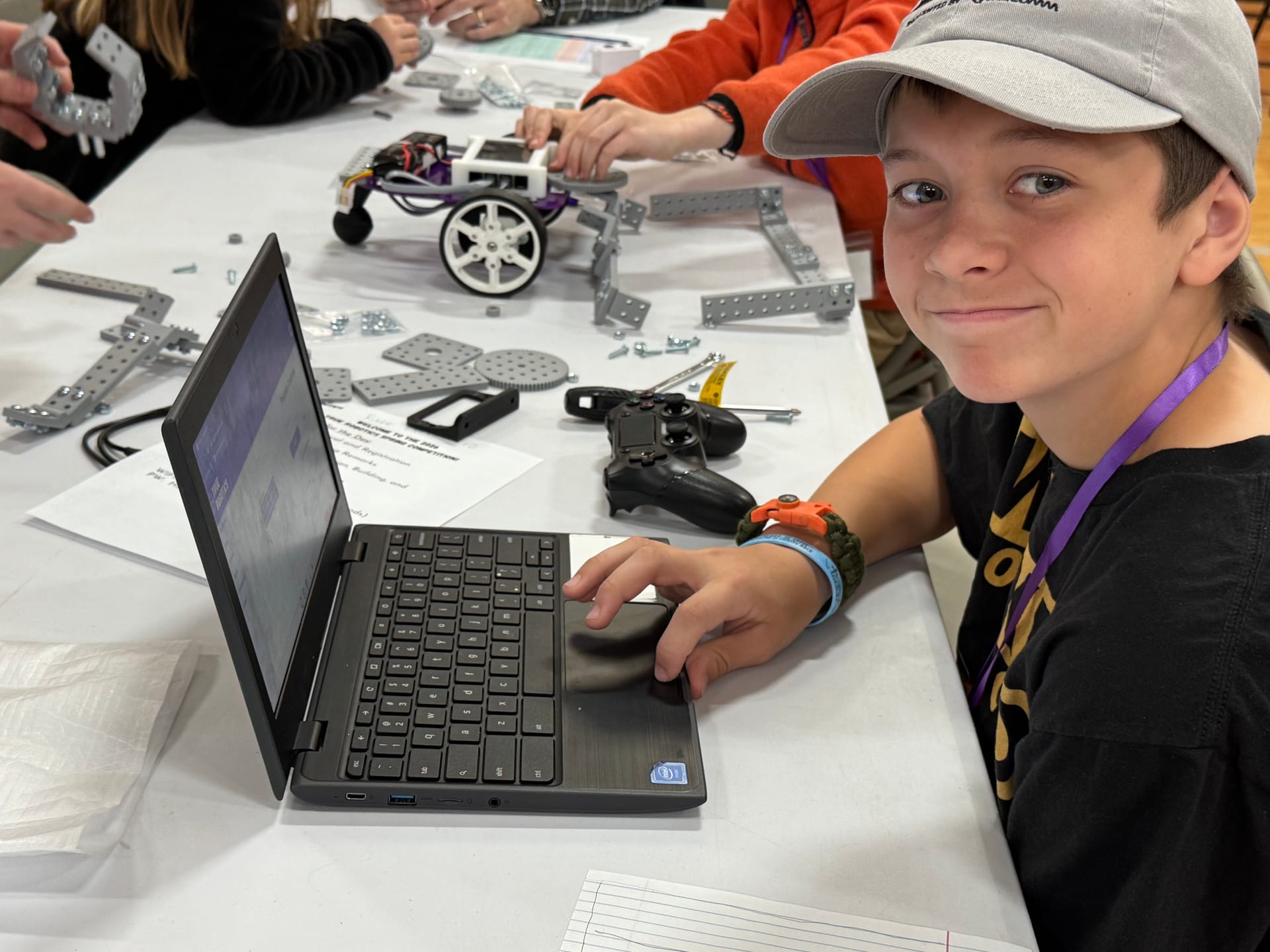 A student looks up from a Chromebook while programming a Pilot robot that sits on the build table beside a game controller.