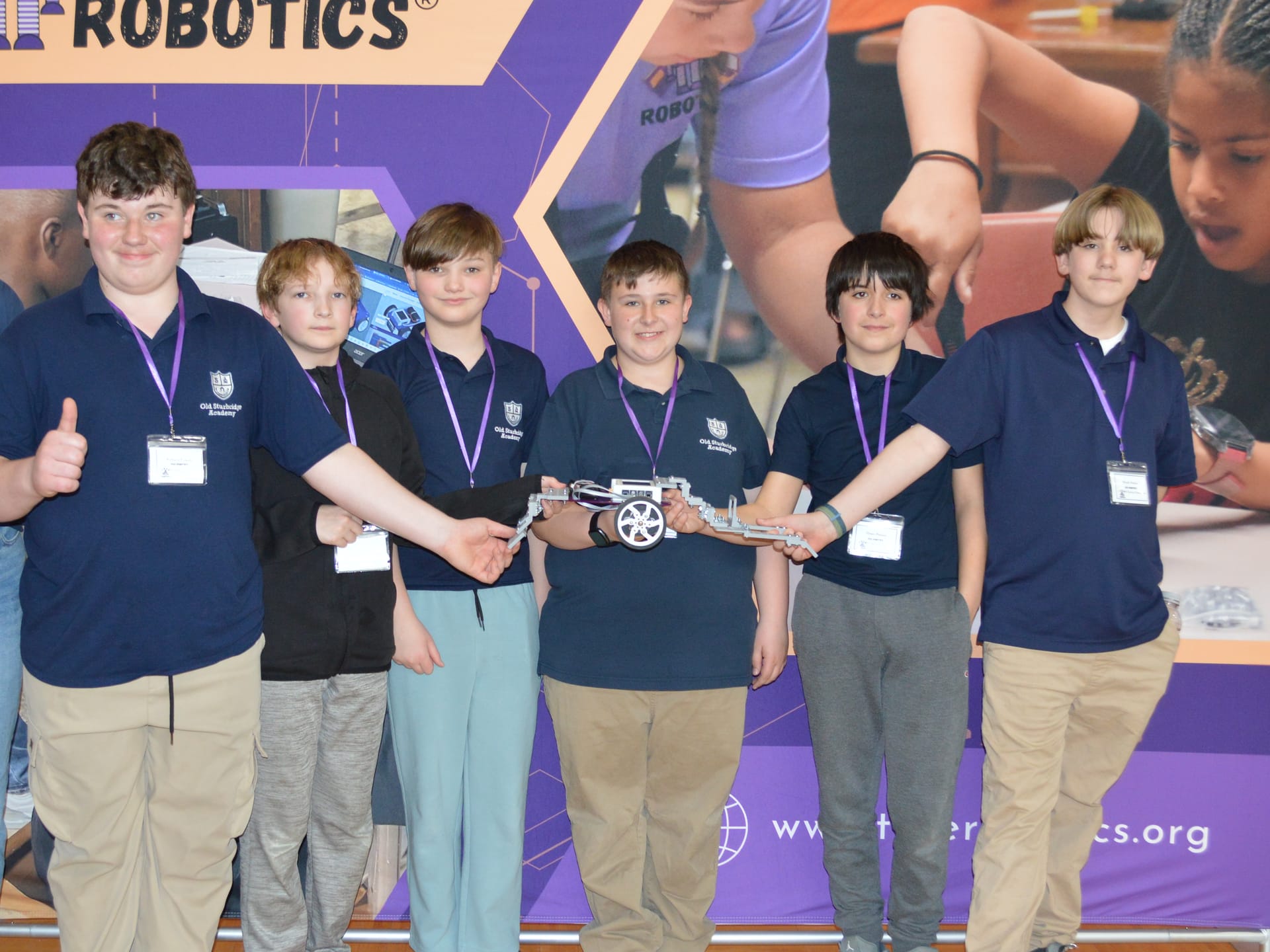 Six students line up at a True Robotics banner holding the robot they built for a competition
