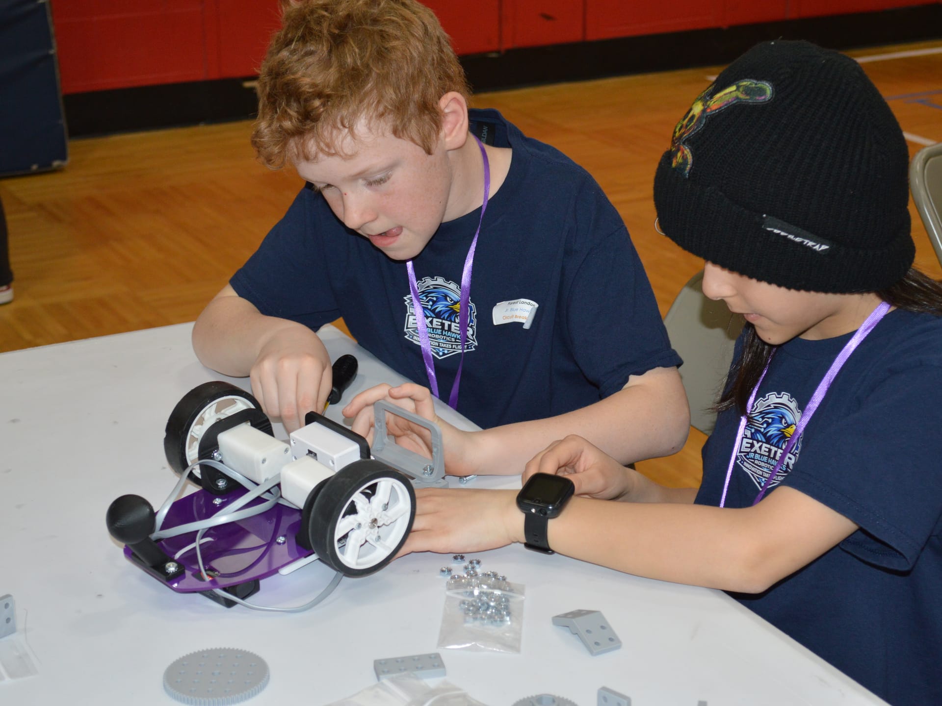 Two students bolt gray FlexForge beams onto a Pilot robot at a competition table