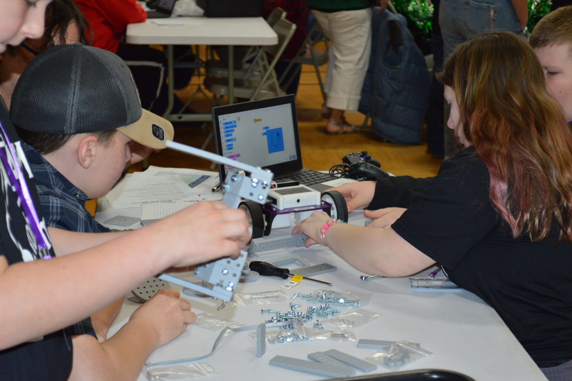 A student holds up a FlexForge arm assembly while a teammate steadies their Pilot robot beside a Chromebook running True Robotics Playground