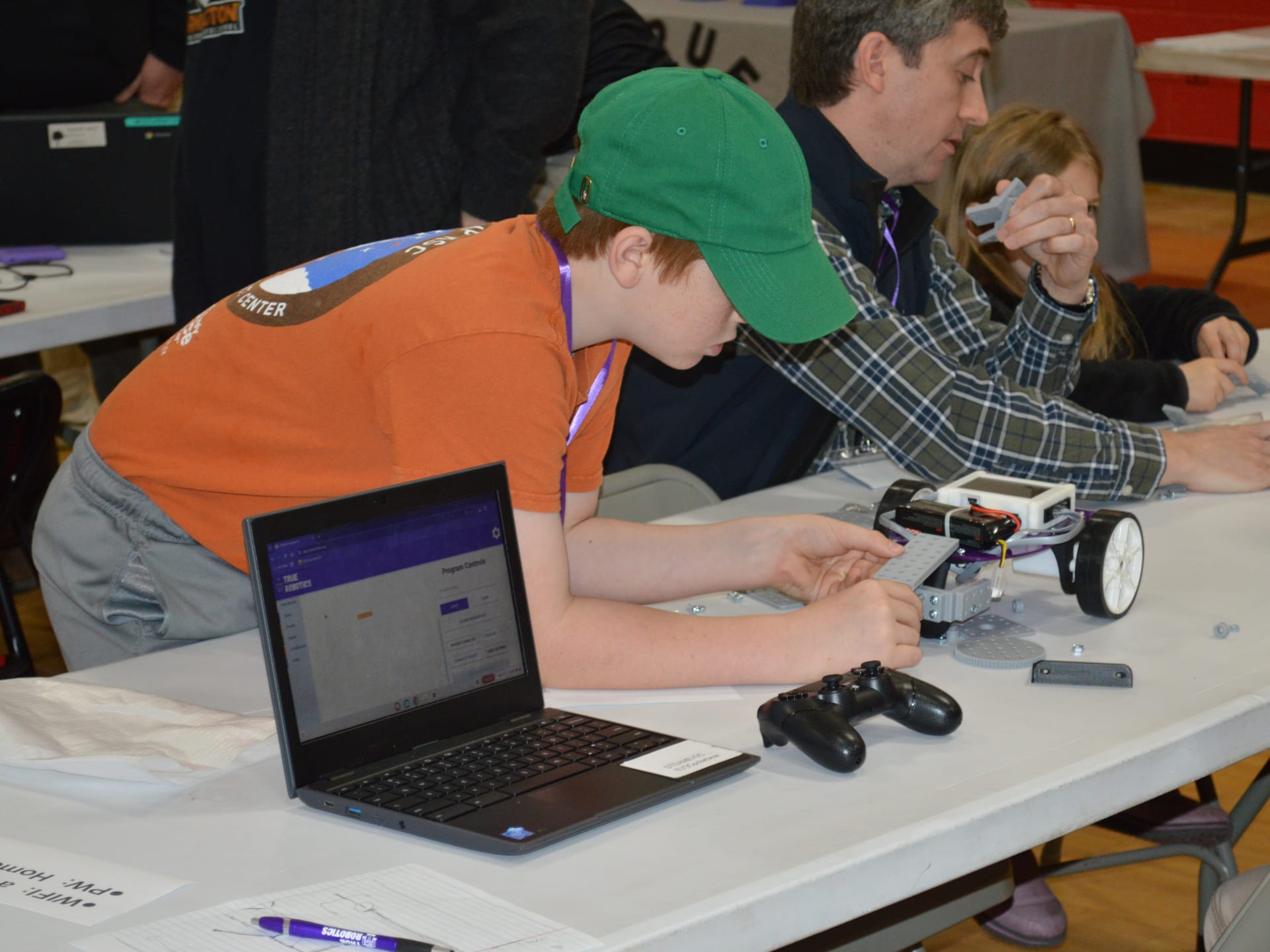 A student fits a FlexForge beam onto a Pilot robot next to a Chromebook running True Robotics Playground and a game controller