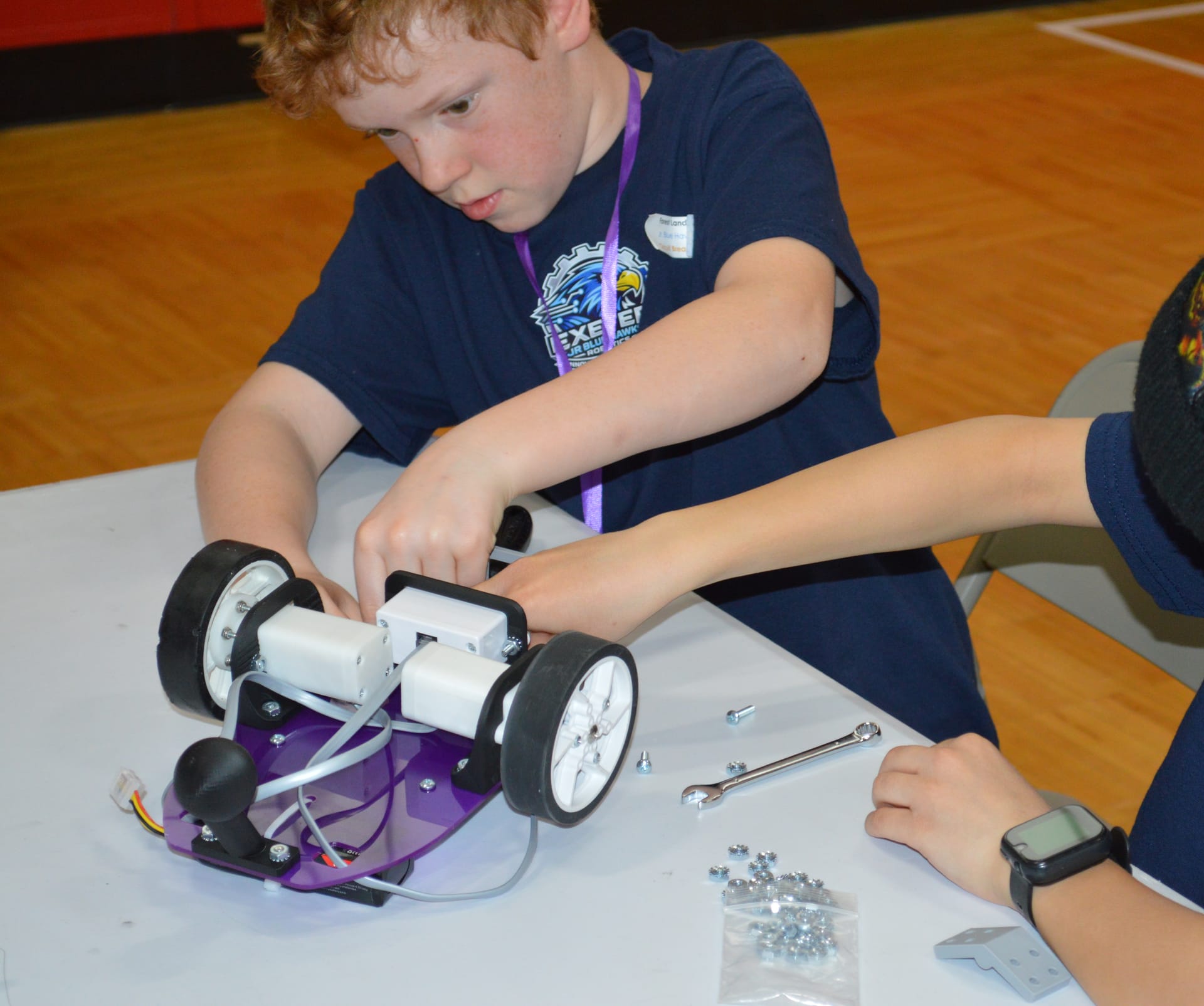 A student bolts white motor mounts onto a purple Pilot robot at a build table with a wrench and loose hardware beside them